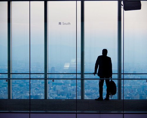 A corporate worker standing up to look towards a distant urban landscape out of a high window pane